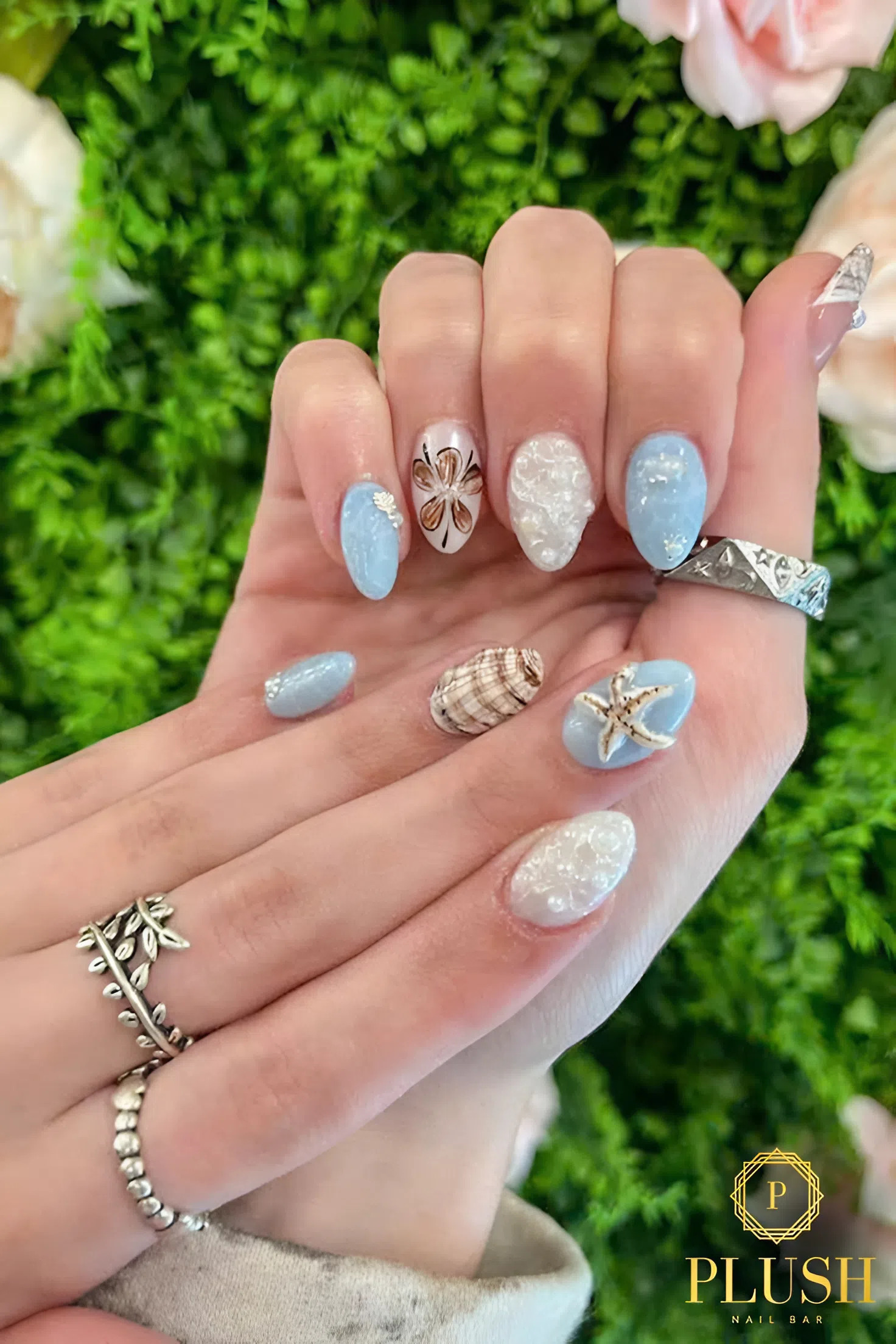Close-up of almond-shaped nails with ocean-inspired designs featuring seashells, pearls, starfish, and soft blue and white polish.