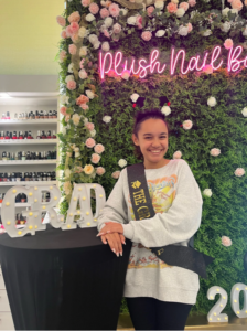 A joyful graduate poses at Plush Nail Bar, showcasing her stylish manicure against a vibrant floral backdrop. With a "Plush Nail Bar" neon sign, decorative flowers, and "Grad 2023" props, this image highlights the perfect place for graduation nails and celebrations. Plush Nail Bar offers elegant manicures for special occasions, helping clients celebrate in style.
