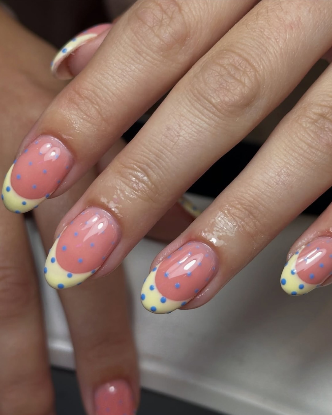 Polka dot nails featuring pastel yellow French tips and bright blue dots on a peachy pink base.
